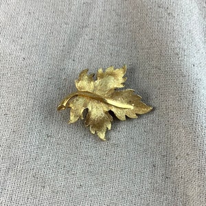 May include: A gold-toned leaf-shaped brooch. The leaf has a detailed design and a textured surface.