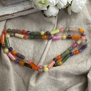 May include: A colorful beaded necklace with a variety of shapes and sizes. The necklace features a mix of cylindrical and spherical beads in shades of orange, pink, green, purple, and yellow. The necklace is displayed on a neutral fabric.