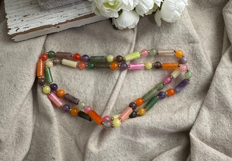 May include: A colorful beaded necklace with a variety of shapes and sizes. The necklace features a mix of cylindrical and spherical beads in shades of orange, pink, green, purple, and yellow. The necklace is displayed on a neutral fabric.