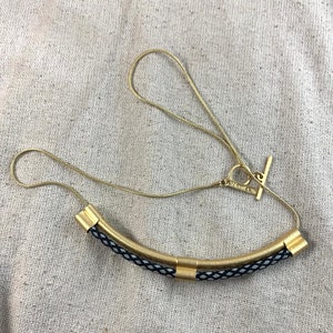 May include: A gold chain necklace with a black and white braided cord and gold accents. The necklace has a toggle clasp.