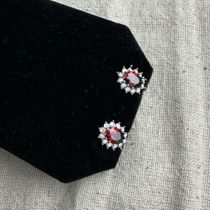 May include: A pair of silver earrings with red oval-shaped gemstones surrounded by smaller clear gemstones.