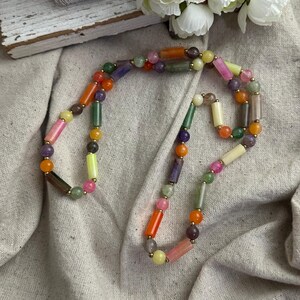 May include: A colorful beaded necklace featuring a variety of shapes and colors, including orange, pink, green, and purple. The necklace has gold-colored accents and is displayed on a neutral-colored fabric.