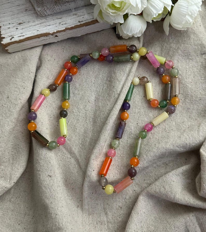 May include: A colorful beaded necklace featuring a variety of shapes and colors, including orange, pink, green, and purple. The necklace has gold-colored accents and is displayed on a neutral-colored fabric.