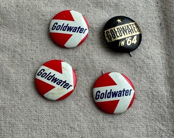 May include: Four vintage political campaign buttons featuring a red, white, and blue arrow pointing to the right with the text "Goldwater" in blue. One button is black with gold text that reads "Goldwater in '64".
