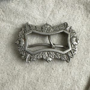 May include: A rectangular silver-toned metal belt buckle featuring an ornate, raised relief border of detailed acanthus leaves and floral motifs. The center of the buckle contains a simple, curved metal prong for securing a belt strap.