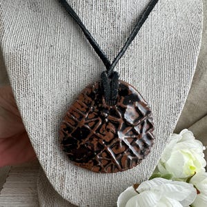 May include: A handcrafted pendant necklace featuring a teardrop-shaped, textured ceramic pendant in shades of brown and black. The pendant is suspended from a black cord. The necklace is displayed on a neutral-toned mannequin.
