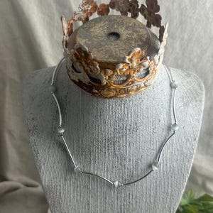 May include: A silver necklace with small, round, light-colored beads and curved bars. The necklace is displayed on a gray bust, with a decorative, rusty-looking crown on top. The crown has a circular base and ornate floral designs.