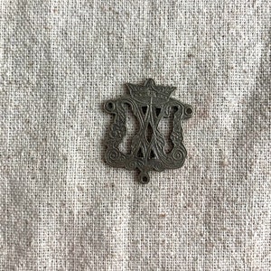 May include: A silver-toned metal pendant with an intricate design featuring a crown and a stylized letter "M".