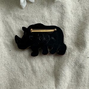May include: A black rhino brooch with a gold-colored pin backing. The rhino is depicted in a side profile, with a textured surface. The brooch is set against a neutral background.