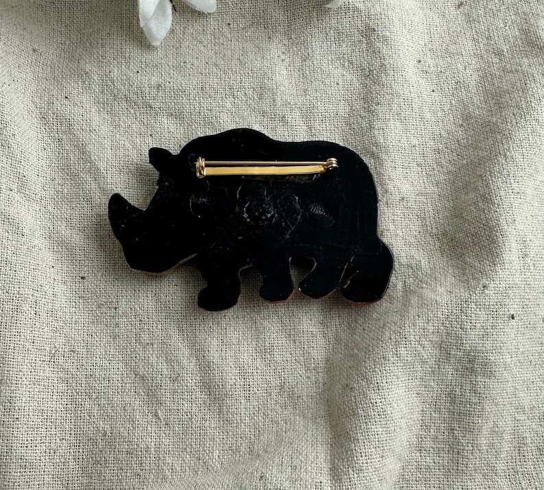 May include: A black rhino brooch with a gold-colored pin backing. The rhino is depicted in a side profile, with a textured surface. The brooch is set against a neutral background.