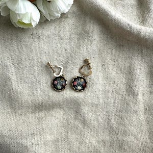 May include: A pair of gold-toned clip-on earrings with a floral design. Each earring features a round, black base with a detailed arrangement of colorful flowers. The earrings are set against a neutral linen background, with white flowers in the upper left corner.