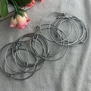 May include: A collection of silver-colored metal hoop earrings with a hinged closure. The earrings are arranged in a pile on a textured, light gray surface. A few pink and peach-colored roses are visible in the background.