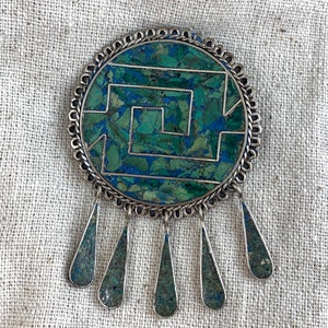 May include: A silver pendant with a round turquoise and silver inlay design. The pendant has a geometric pattern and five teardrop-shaped turquoise and silver drops hanging from the bottom.
