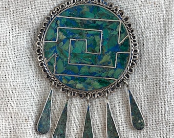 May include: A silver pendant with a round turquoise and silver inlay design. The pendant has a geometric pattern and five teardrop-shaped turquoise and silver drops hanging from the bottom.