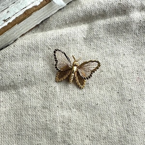 May include: A gold-toned butterfly brooch with detailed wing patterns and a small, clear gemstone. The brooch is set against a neutral linen background, highlighting its intricate design and vintage aesthetic.