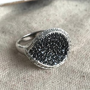 May include: A silver ring with a black and white oval-shaped stone. The stone is set in a pave setting and is surrounded by a border of smaller white stones.