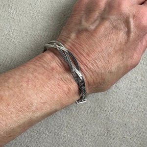 May include: A silver chain bracelet with a woven design. The bracelet is on a person's wrist.