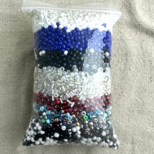 May include: A clear plastic bag filled with various colors of seed beads. The beads are arranged in layers, with white, blue, black, silver, red, and multi-colored beads.