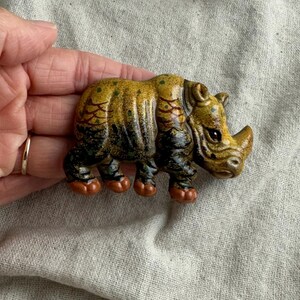May include: A detailed, hand-painted ceramic rhinoceros figurine. The rhino features a textured, yellow and brown body with black accents on its legs and a small horn. The figurine is held in a hand.