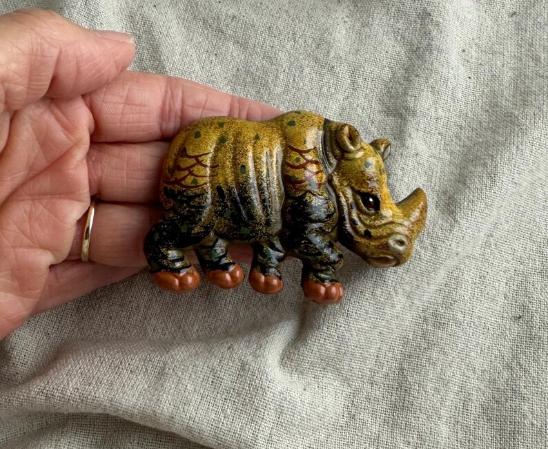 May include: A detailed, hand-painted ceramic rhinoceros figurine. The rhino features a textured, yellow and brown body with black accents on its legs and a small horn. The figurine is held in a hand.