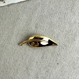 May include: A gold-toned leaf-shaped brooch with a small square logo on the back.