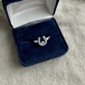May include: A silver ring with a horseshoe design, encrusted with small, clear gemstones. The ring is displayed inside a dark blue velvet ring box with a white satin interior. The box is open, revealing the ring.