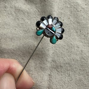 May include: A silver stick pin featuring a fan-shaped inlay design. The decorative head contains inlaid segments of black onyx, white mother-of-pearl, red coral, and two teardrop-shaped turquoise stones arranged in a radial pattern.