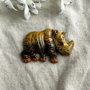May include: A ceramic rhino figurine with a detailed design. The rhino is primarily yellow and brown with red and green accents. The figurine has a textured appearance and is resting on a neutral-colored surface.