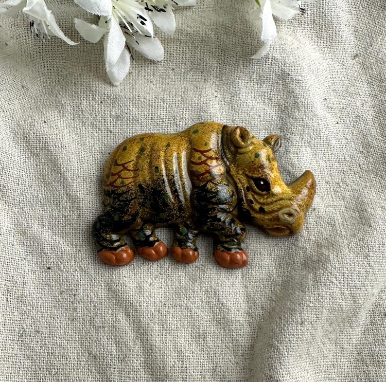 May include: A ceramic rhino figurine with a detailed design. The rhino is primarily yellow and brown with red and green accents. The figurine has a textured appearance and is resting on a neutral-colored surface.
