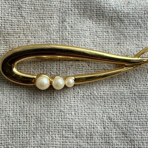 May include: A gold-toned brooch with a flowing, open design. The brooch features three small, round, off-white pearls set along the curve. The pin is set against a textured, light gray fabric.