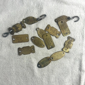 May include: A collection of vintage brass tags with various shapes and sizes, including oval, rectangular, and irregular designs. The tags feature stamped text and numbers, some with attached metal hooks or loops. The tags are arranged on a light-colored fabric.