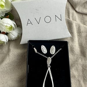 May include: A silver-toned jewelry set, including a necklace and matching earrings, presented in a black box. The necklace features a teardrop pendant adorned with sparkling crystals. The earrings are oval-shaped, also embellished with crystals. The box is from the brand AVON.