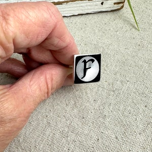 May include: A silver cufflink with a black square design featuring a white circle with a black letter "F" in a stylized font.