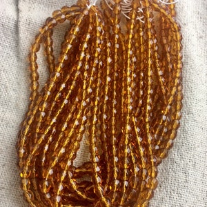 May include: A bundle of amber glass beads, each bead is faceted and has a round shape. The beads are strung together on a white thread.