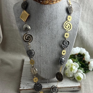 May include: A long necklace featuring a mix of gold and silver-toned metal charms, including hearts, spirals, squares, and floral shapes, connected by thin metal links. The necklace is displayed on a gray bust with a decorative metal crown.