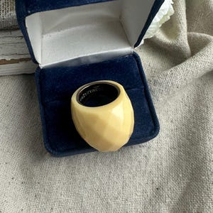 May include: A faceted, ivory-colored ring with a silver interior sits in a blue velvet ring box. The ring has a geometric design and a polished finish, showcasing a modern aesthetic. The ring is a statement piece.