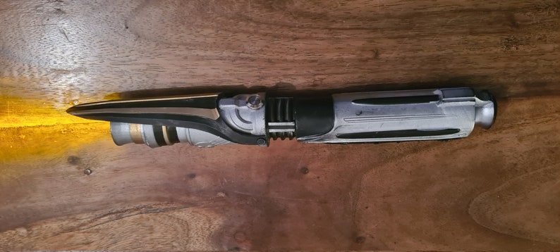 May include: A black, silver, and gold lightsaber prop with a textured handle and a black blade.