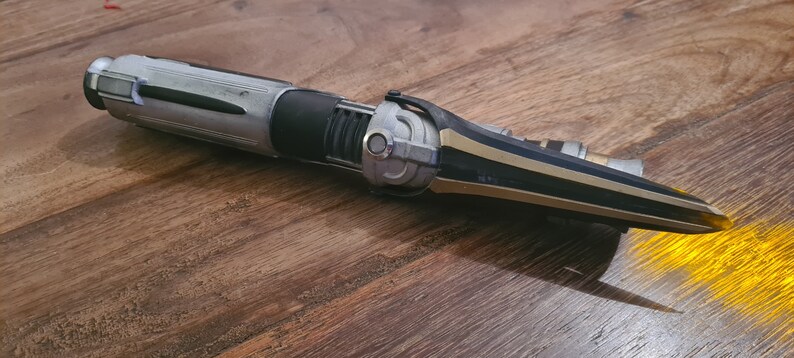 May include: A black and silver lightsaber with a gold accent. The lightsaber is in a deactivated state and has a sharp, pointed blade.
