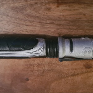 May include: A silver and black lightsaber with a textured grip and a gold accent at the end.
