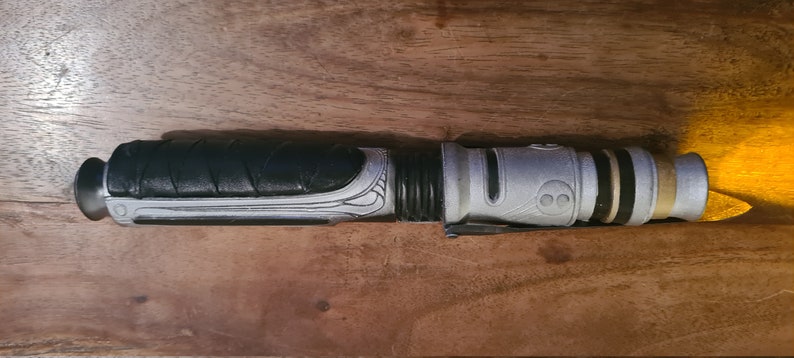 May include: A silver and black lightsaber with a textured grip and a gold accent at the end.