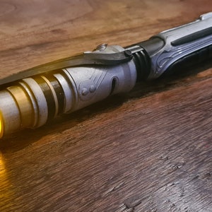 May include: A silver and black lightsaber with a yellow blade. The lightsaber is on a wooden surface.