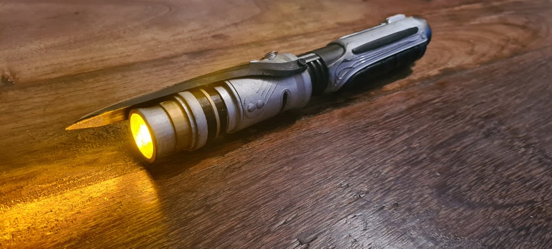 May include: A silver and black lightsaber with a yellow blade. The lightsaber is on a wooden surface.