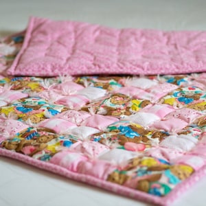 Puede incluir: Una manta de bebé acolchada rosa y blanca con un diseño de patchwork con ositos de peluche. La manta tiene un borde rizado y está hecha de tela suave y mullida.