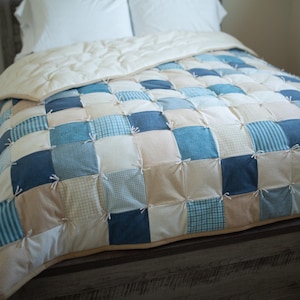 May include: A patchwork quilt with squares of blue, white, and beige fabric. The quilt is tied with white string and is on a bed with a wooden headboard.