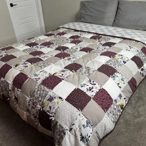 Puede incluir: Un edredón hecho a mano con un diseño de patchwork. El edredón presenta cuadrados en tonos burdeos, crema, gris y estampados florales. El edredón está sobre una cama con almohadas grises y una sábana superior a cuadros.