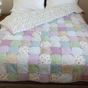 Puede incluir: Una colcha de patchwork en colores pastel, incluyendo lavanda, verde y crema, cubre una cama. La colcha presenta bloques cuadrados atados con lazos de lavanda. También se ve una funda nórdica estampada a juego y almohadas blancas.