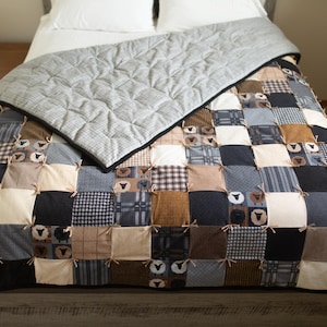 May include: A patchwork quilt with a grey top layer and a patterned bottom layer. The bottom layer features squares of various fabrics in shades of black, brown, beige, and blue, some with sheep designs. The quilt is on a bed with white pillows.