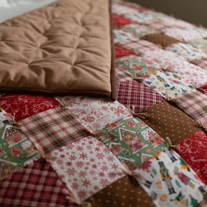 May include: A patchwork quilt featuring a variety of holiday-themed fabric squares in red, green, white, and brown. The patterns include nutcrackers, gingerbread houses, snowflakes, and plaid designs. The reverse side is a solid tan fabric.