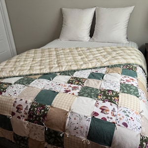 May include: A patchwork quilt featuring a variety of square fabric blocks in shades of green, beige, and white. The design includes patterns of mushrooms, botanical illustrations, and solid textures, finished with tied seams and a plaid top edge.
