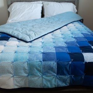 Peut inclure: Une courtepointe faite à la main dans des tons de bleu, du clair au foncé, disposée en patchwork. La courtepointe est sur un lit avec des oreillers blancs et un drap supérieur bleu clair. Les carrés sont noués avec du fil bleu.